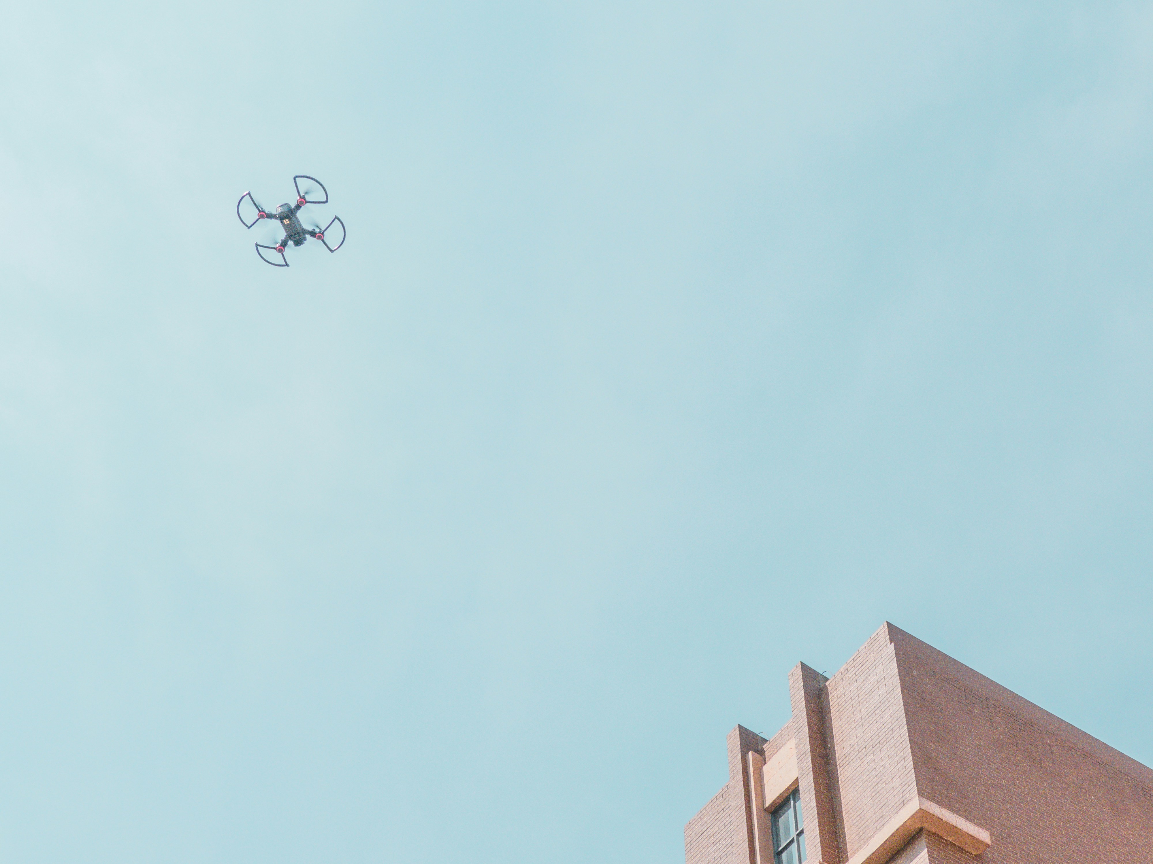 Drone flying near a rooftop against a pale blue sky beside a brick building
