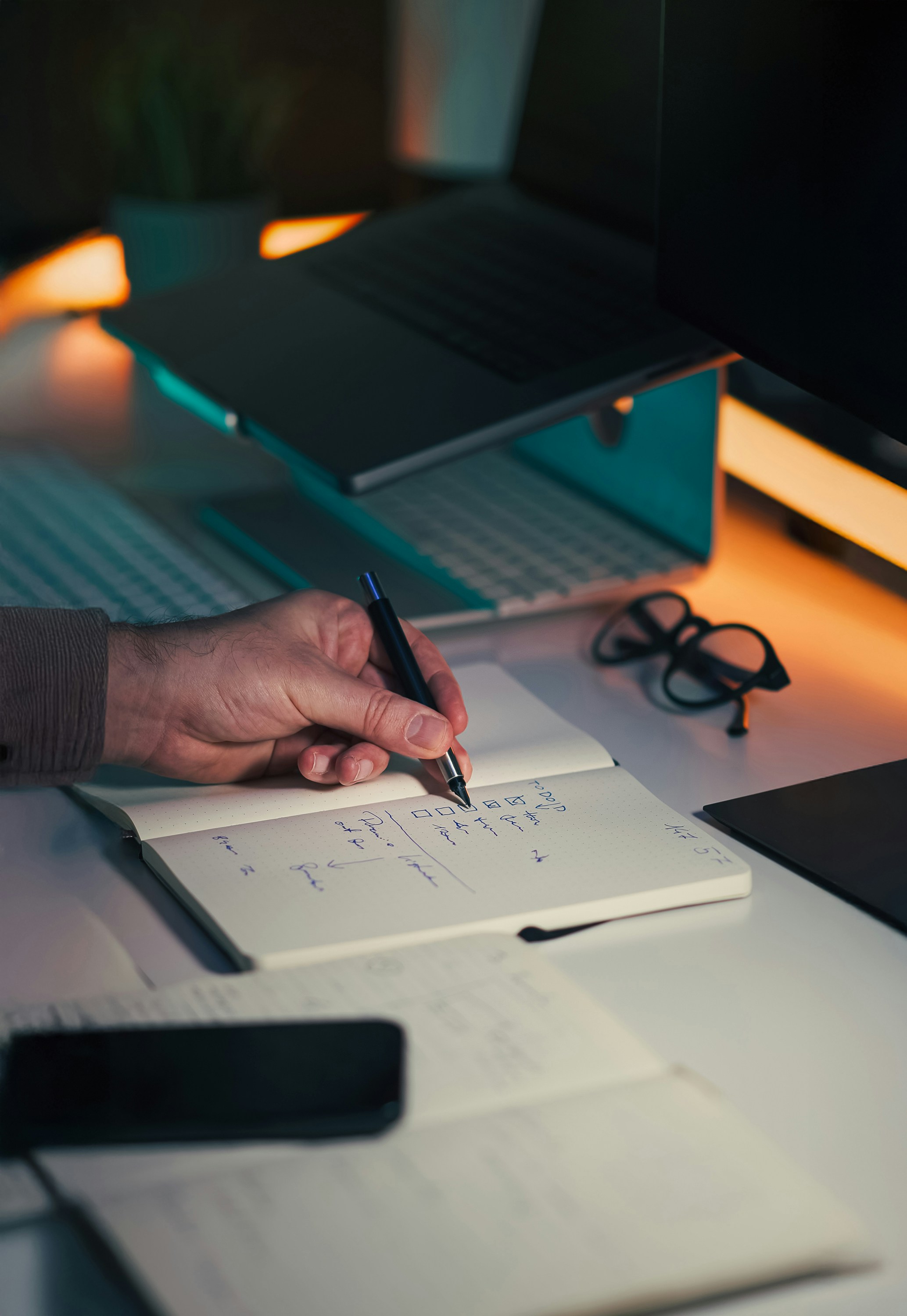 Close-up of a hand writing notes in a notebook at a desk with a laptop and glasses.