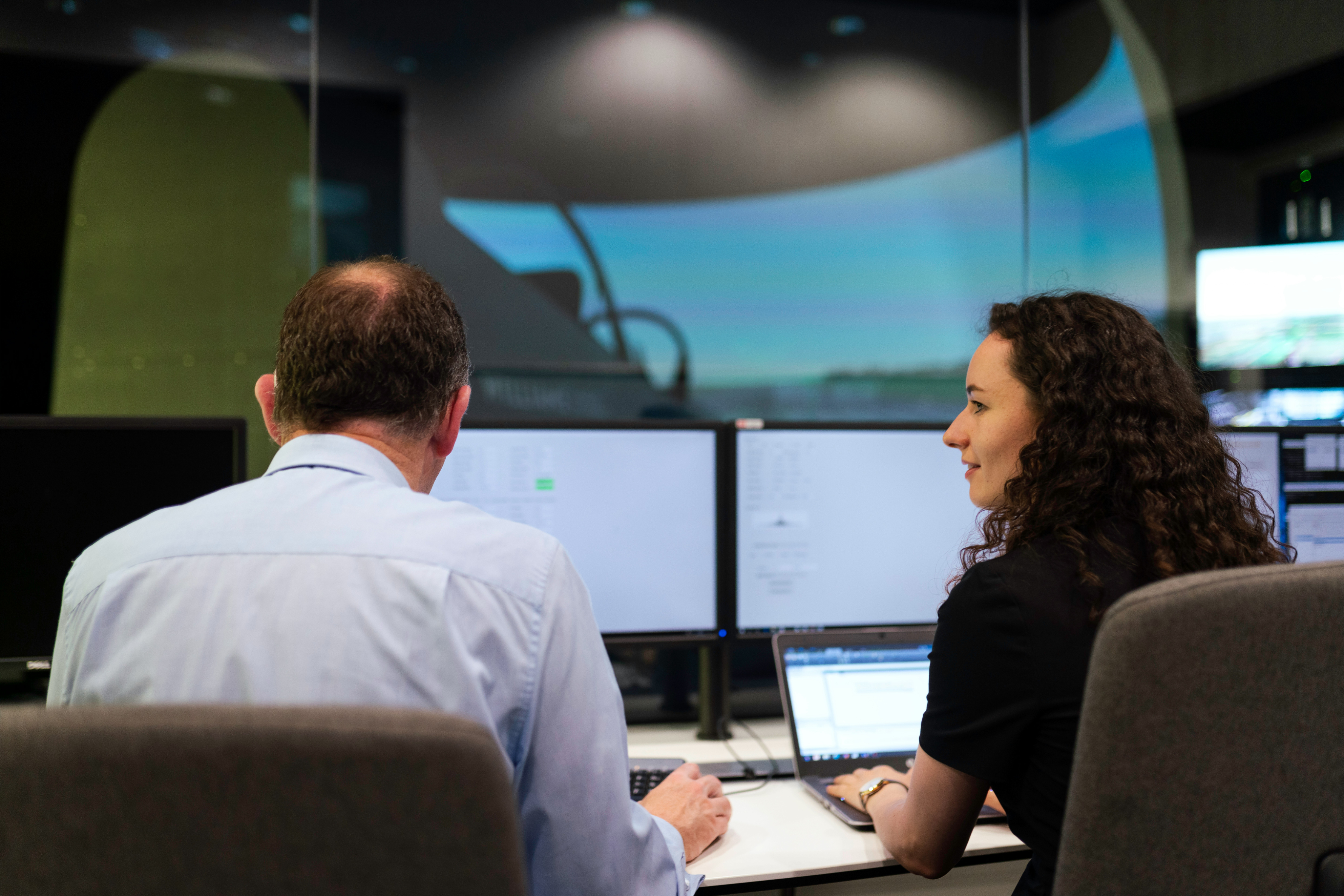 Man and woman reviewing multiple screens and a laptop in a control room