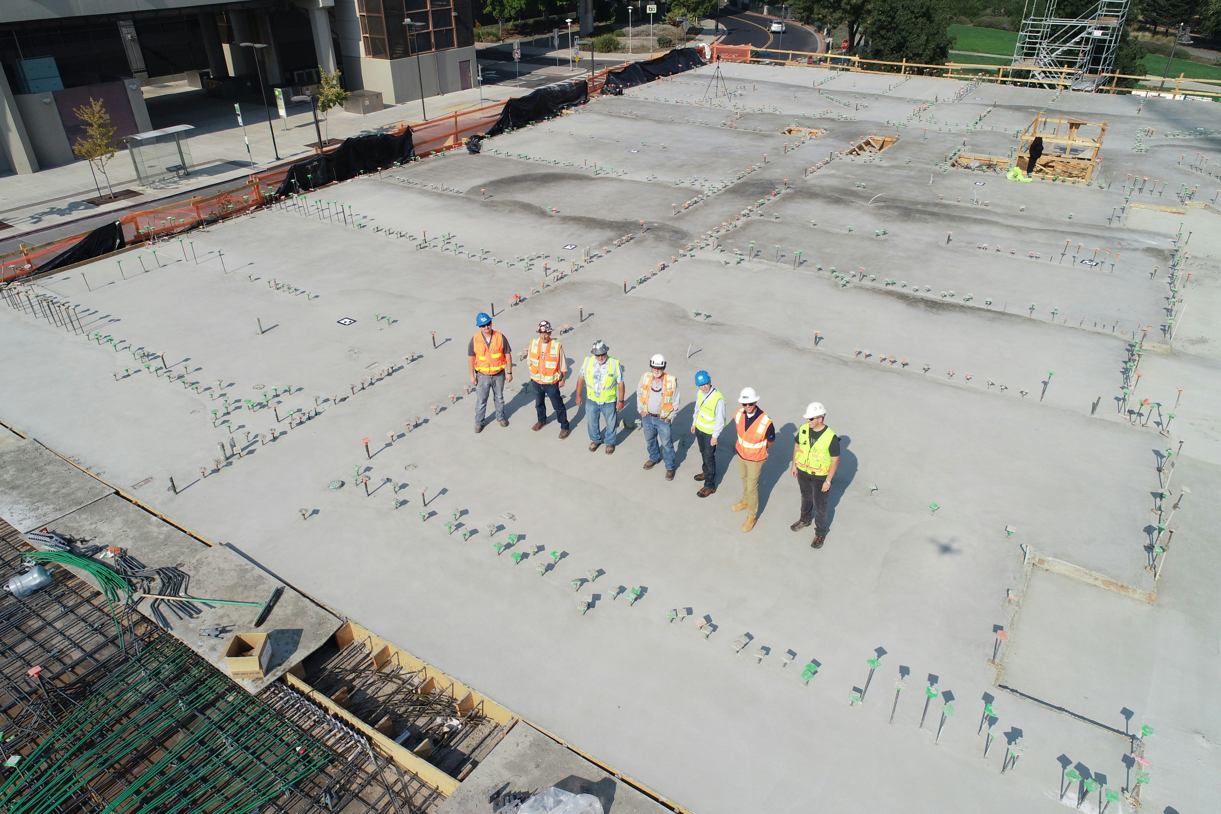 Aerial view of seven construction workers standing on a concrete slab, drone shadow visible